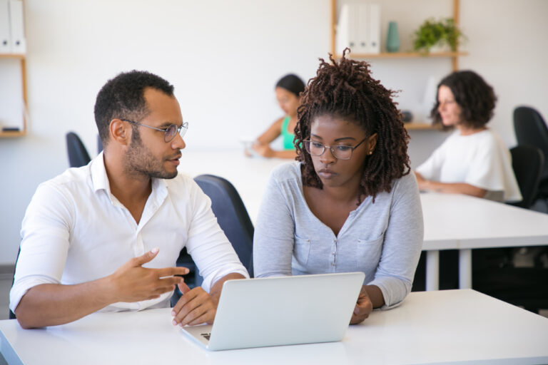 Business professional explaining project details to colleague. Man and woman  sitting at table in office, using laptop, looking at screen and talking. Consulting concept
